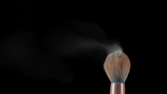 Flow of air blows fine powder particles off makeup brush. Crushed dust of beige powder scatters in space against black background. Great makeup brushes close up. Powder brush. Slow motion
