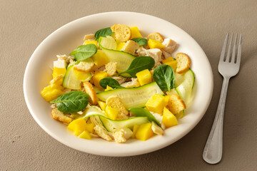 Plate with salad and fork close-up on a brown background, salad of cucumber and chicken breast, mozzarella and croutons with sweet mango pieces and spinach