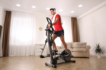 Man with headphones and towel using modern elliptical machine at home