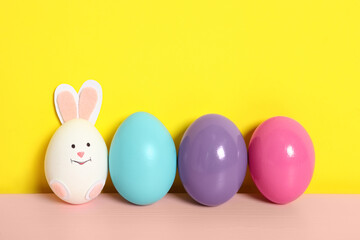 Bright eggs and white one as cute bunny on pink wooden table against yellow background, space for text. Easter celebration