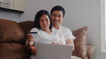 Young Asian Pregnant couple online shopping at home. Mom and Dad feeling happy using laptop technology and credit card buying baby product while lying on sofa in living room at home concept.