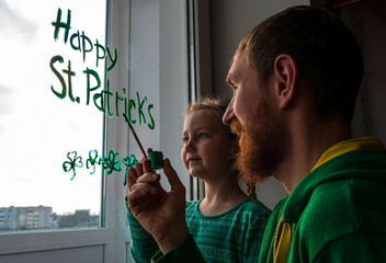 Drawing St. Patrick's Day Father with daughter painting green three-leaved shamrocks indoor, festive home decoration,family leisure. Drawing clover leaves on window glass. Stay home concept New normal