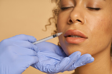 Close up shot of cosmetic injection to the lips of pretty young woman isolated over beige background