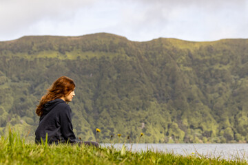 Fototapeta na wymiar Girl sitting in grass, by Sete Cidades lake, enjoying the landscape, peacefully.