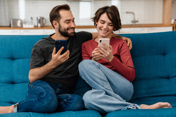 Happy young couple smiling and using mobile phones while sitting on couch