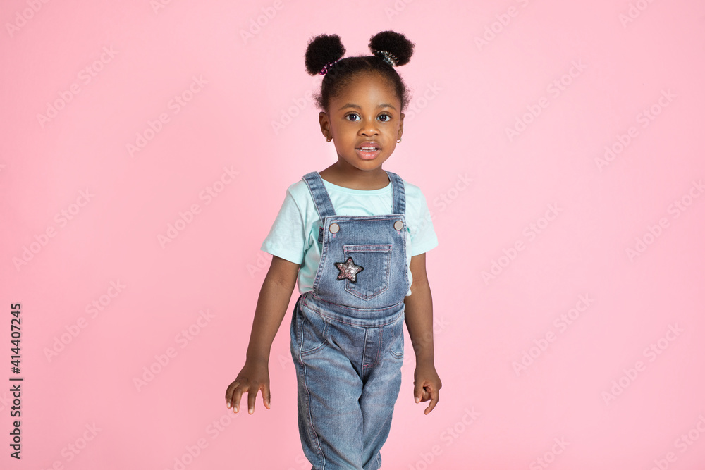 Poster Cute little African preschool girl, wearing casual jeans overalls and t-shirt, posing to camera on isolated pink studio background. People, kids, studio portraits