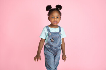 Cute little African preschool girl, wearing casual jeans overalls and t-shirt, posing to camera on isolated pink studio background. People, kids, studio portraits