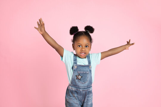 Half length portrait of cute little african kid girl in jeans overalls, isolated over pink background, standing with her arms outstretched