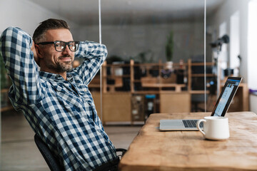 Pleased unshaven man in eyeglasses resting while working with laptop