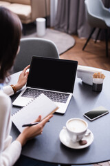 Fototapeta premium cropped view of freelancer holding notebook near laptop with blank screen, blurred foreground