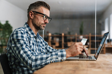 Focused unshaven man using credit card while working with laptop