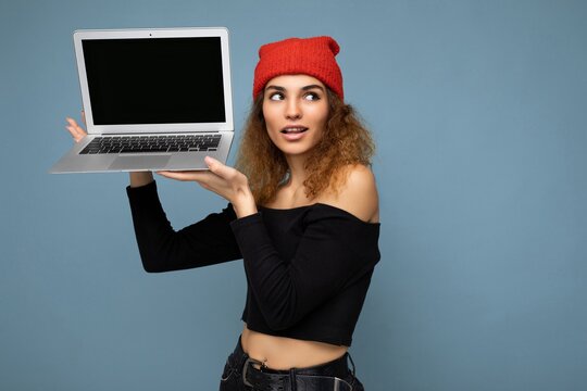 Close-up portrait of beautiful funny charming self-confident dark blond curly young woman holding laptop computer looking up wearing black crop top and red and orange do-rag isolated over light blue