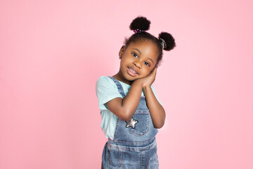 Close up portrait of little adorable African child girl in jeans overalls, having fun and looking at camera over pink background. Copy space