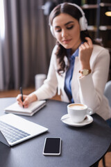 freelancer in headphones writing in notebook near laptop and smartphone with blank screen, blurred background