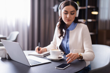 young freelancer in headphones working in restaurant near laptop and cellphone with blank screen, blurred background