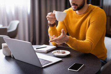 cropped view of arabian freelancer pointing at laptop while holding cup of coffee