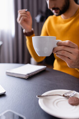 cropped view of arabian teleworker holding cup of copy near notebook in restaurant, blurred background