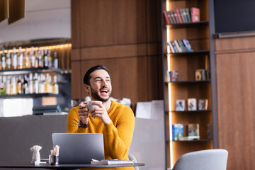 excited arabian freelancer laughing while holding cup of coffee in restaurant