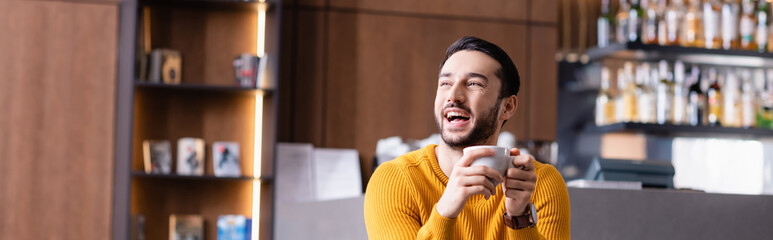 excited arabian man laughing while holding cup of coffee and looking away in restaurant, banner