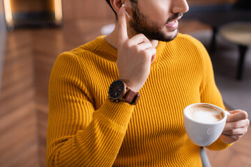 cropped view of arabian man adjusting earphone while holding cup of coffee in restaurant