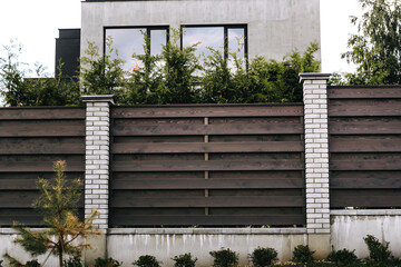 Horizontal sections of brown wooden boards fence and white brick pillars. Live plantings. Green thuja, bushes and pine. Territory landscaping. Capital fencing. Production of fence for a country house