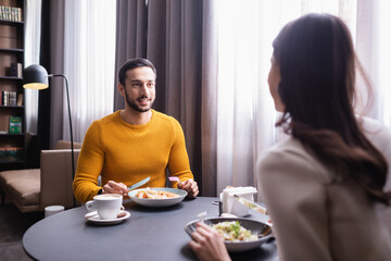 Arabian man smiling at girlfriend on blurred foreground near food and coffee in restaurant