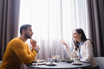 Smiling woman talking to arabian boyfriend near food and coffee in restaurant