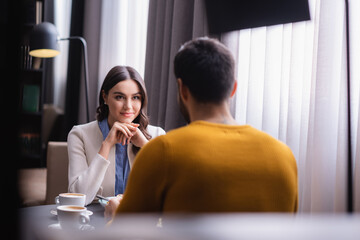 Young woman looking at boyfriend near cups of cappuccino on blurred foreground in restaurant