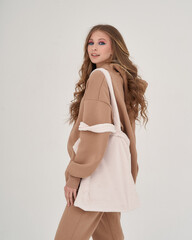 Sporty teenager girl with long blonde wavy hair in beige pants and hoodie standing against white background and holding textile bag