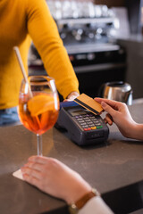 Cropped view of woman holding credit card near payment terminal, bartender and cocktail on blurred foreground