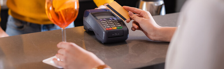 Cropped view of woman paying with credit card near cocktail and barman on blurred background, banner