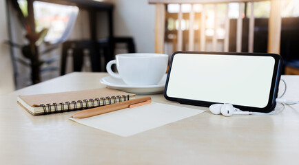 Close-up, blank screen mobile with headphones and book, pen, coffee cup on wooden table