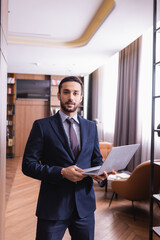 Fototapeta na wymiar Young arabian businessman holding laptop and looking at camera in restaurant