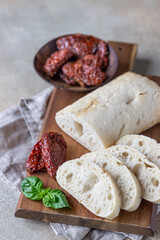 Freshly baked ciabatta bread on wooden plate with sun-dried tomatoes and basil, concrete background.