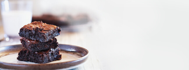 Banner of fresh made homemade fudgy brownies stacked on a saucer over a white rustic wooden table. Extreme shallow depth of field with blurred background and a glass of milk.