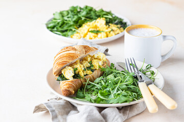 Delicious croissant sandwich with scrambled spinach eggs served with arugula on plate and coffee cup, light concrete background.