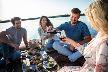 Group of friends having fun on picnic near a lake, sitting on pier eating and drinking wine.