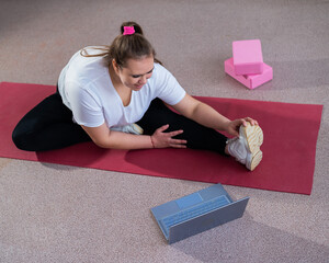 Young caucasian fat woman doing bends on a sports mat and watching a training video on a laptop. A...