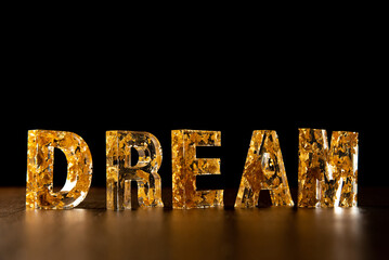 Acrylic letters with gold leaves forming the word dream on wooden surface, black background, selective focus.