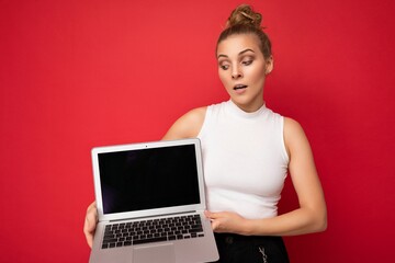 beautiful blond young woman with gathered hair looking down at netbook keyboard holding computer laptop with empty monitor screen with mock up and copy space wearing white t-shirt isolated on red wall