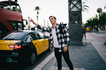 Young caucasian male traveler with backpack hailing on road in megalopolis standing on street, positive casual dressed man 20s tourist stopping cab raising hand gesture for taking transport in city