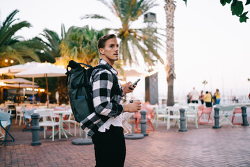 Serious caucasian 20s man traveler with backpack looking for direction in city using mobile phone...