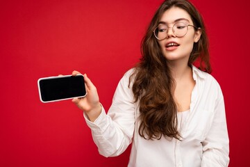 charming young brunet woman wearing white shirt and optical glasses isolated over red background holding in hand and showing mobile phone with empty screen for mockup looking at gadjet screen