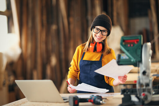 Smiling craftswoman working with laptop and papers in joinery workshop