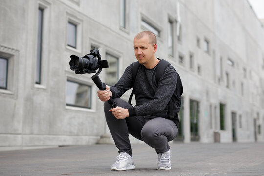 videography, filmmaking and creativity concept - professional male videographer shooting video using modern dslr camera on 3-axis gimbal over grey concrete building