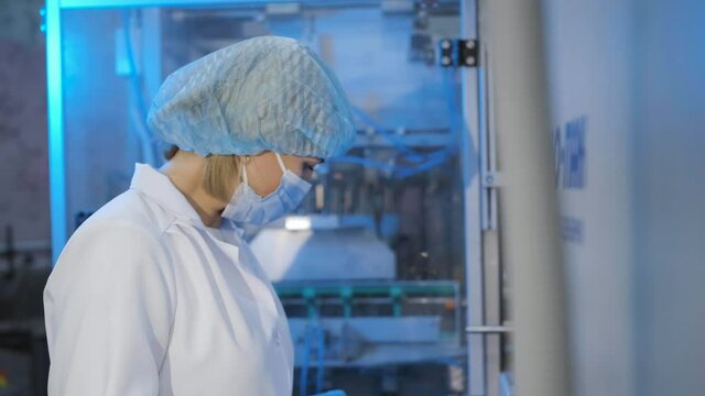 A middle-aged woman operator in a white coat, disposable cap, medical mask and latex gloves near production equipment presses buttons.