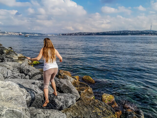 Young slender woman runs on huge stones on Bosphorus embankment on the backdrop of Istanbul panorama (Turkey) - a view from the back. Adult barefoot long-haired girl jumping on boulders by the sea