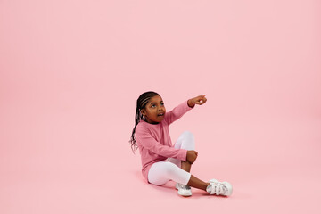 Pointing, showing. Childhood and dream about big and famous future. Pretty little girl isolated on coral pink studio background. Dreams, imagination, education, facial expression, emotions concept.