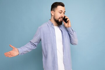 Photo shot of good looking young brunet bearded man wearing casual blue shirt and white t-shirt poising isolated on blue background with empty space holding in hand and communicating talking on mobile