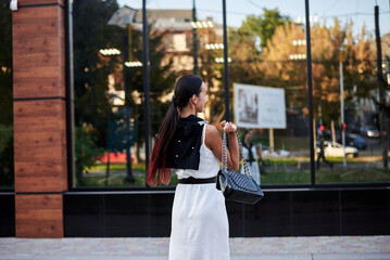 Young brunette girl with red pony tail, wearing stylish white silk dress,running jumping in front of glass building, holding phone, smiling.Business woman on lunch break.Romantic female urban portrait